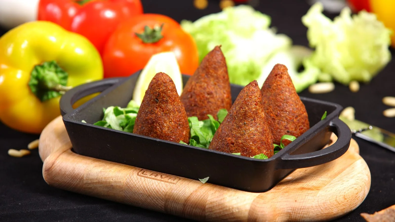 Appetizing kibbeh served in a cast iron dish on a wooden board, surrounded by fresh vegetables.