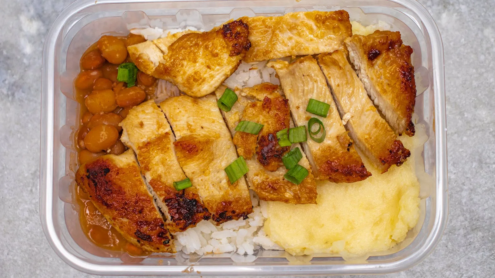 Top view of a tasty home-cooked meal with grilled chicken, beans, and rice in a bento box.