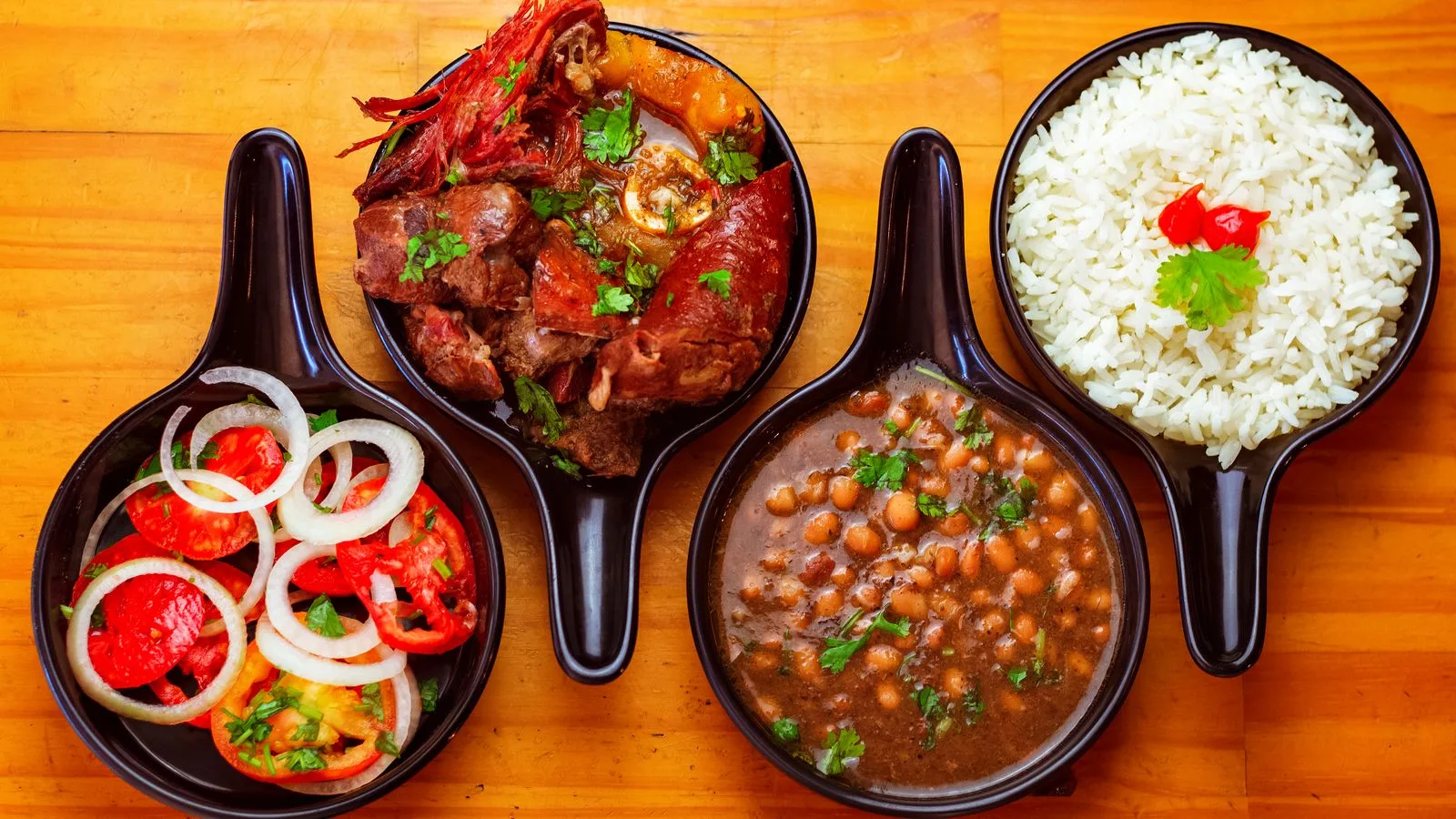 Top view of traditional Brazilian dishes with rice, beans, beef, and fresh salad served in black bowls.