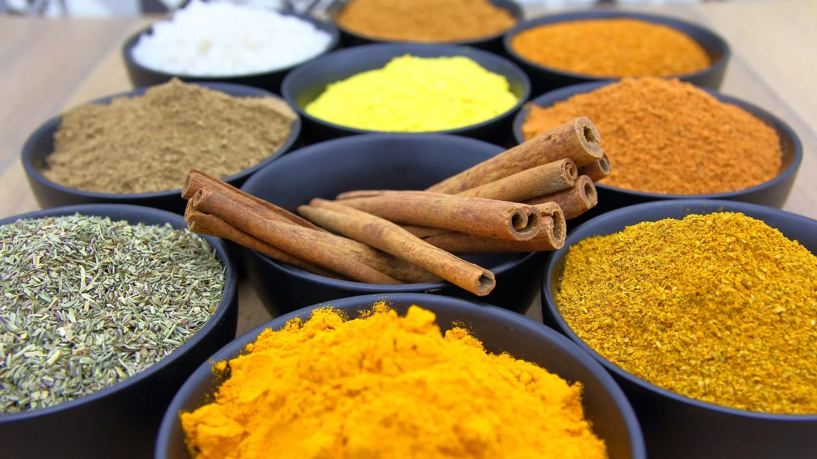 Colorful assortment of spices in bowls with cinnamon sticks on a wooden surface.