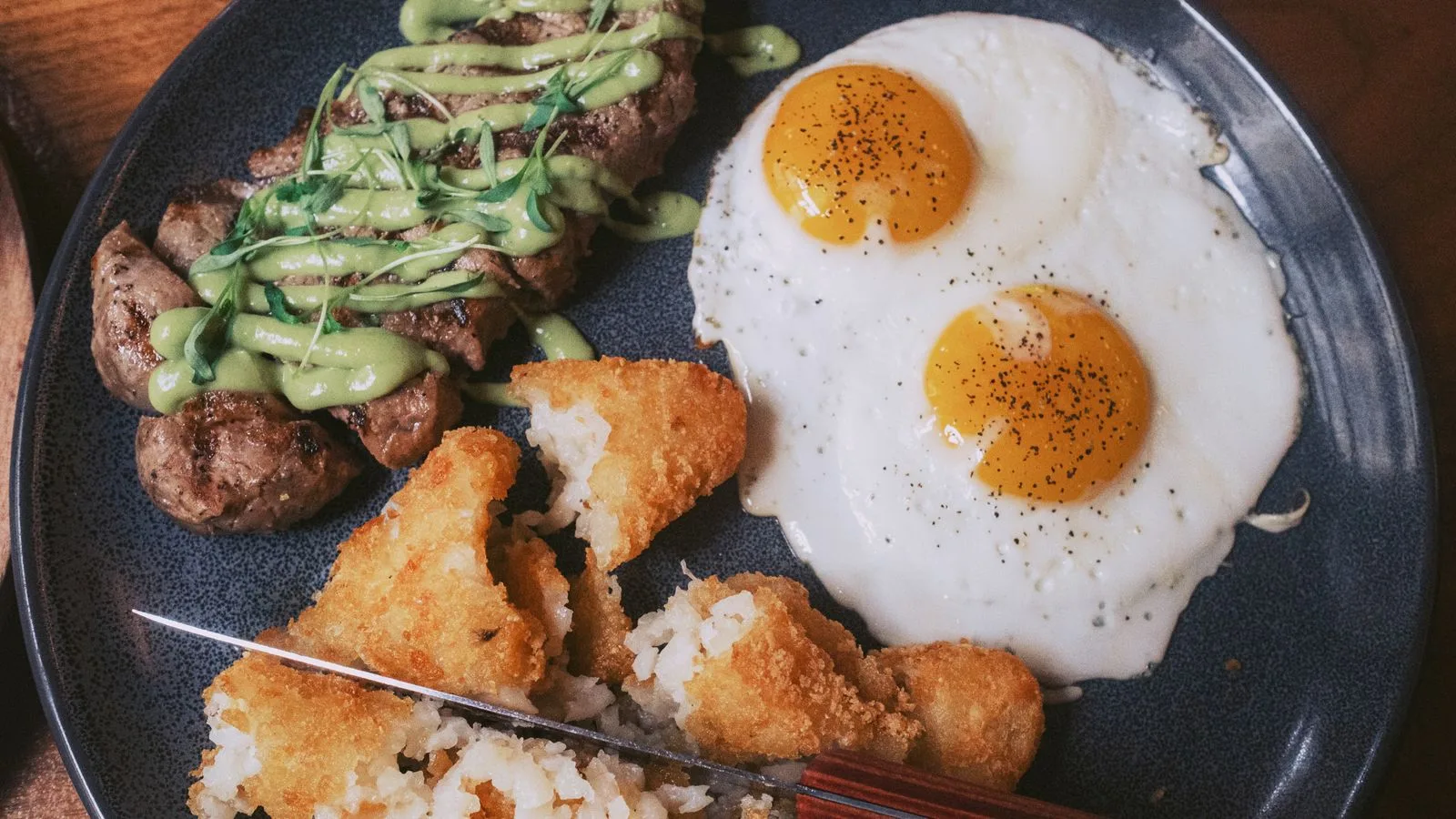 Delicious breakfast plate featuring steak with sauce, fried eggs, and crispy potato cakes.