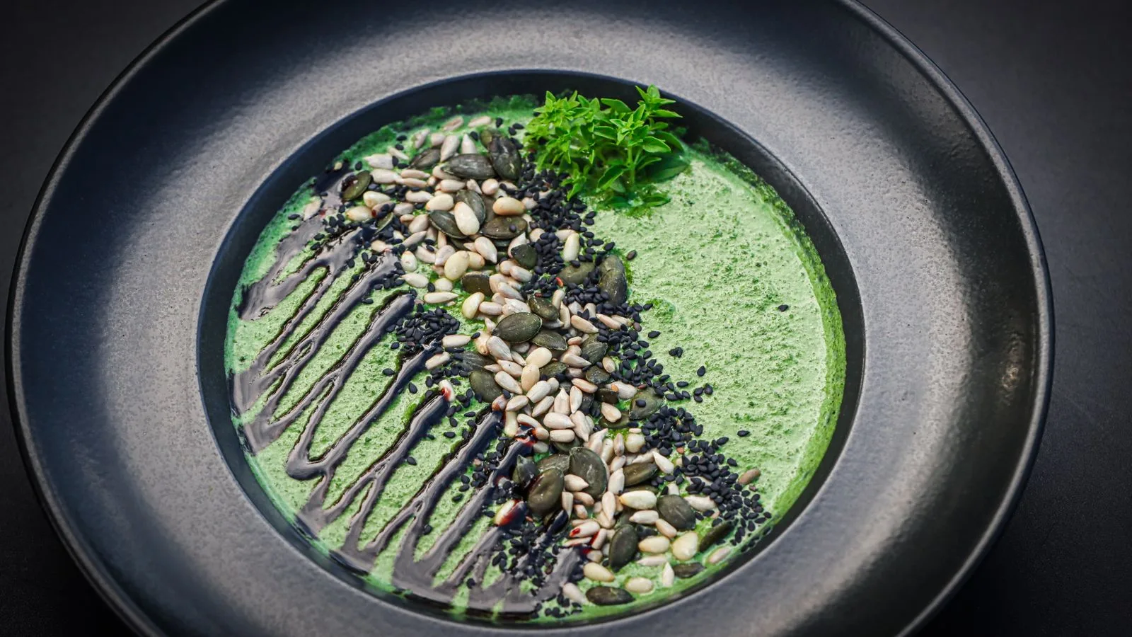 A gourmet green soup served in a black bowl, topped with mixed seeds and drizzled sauce.