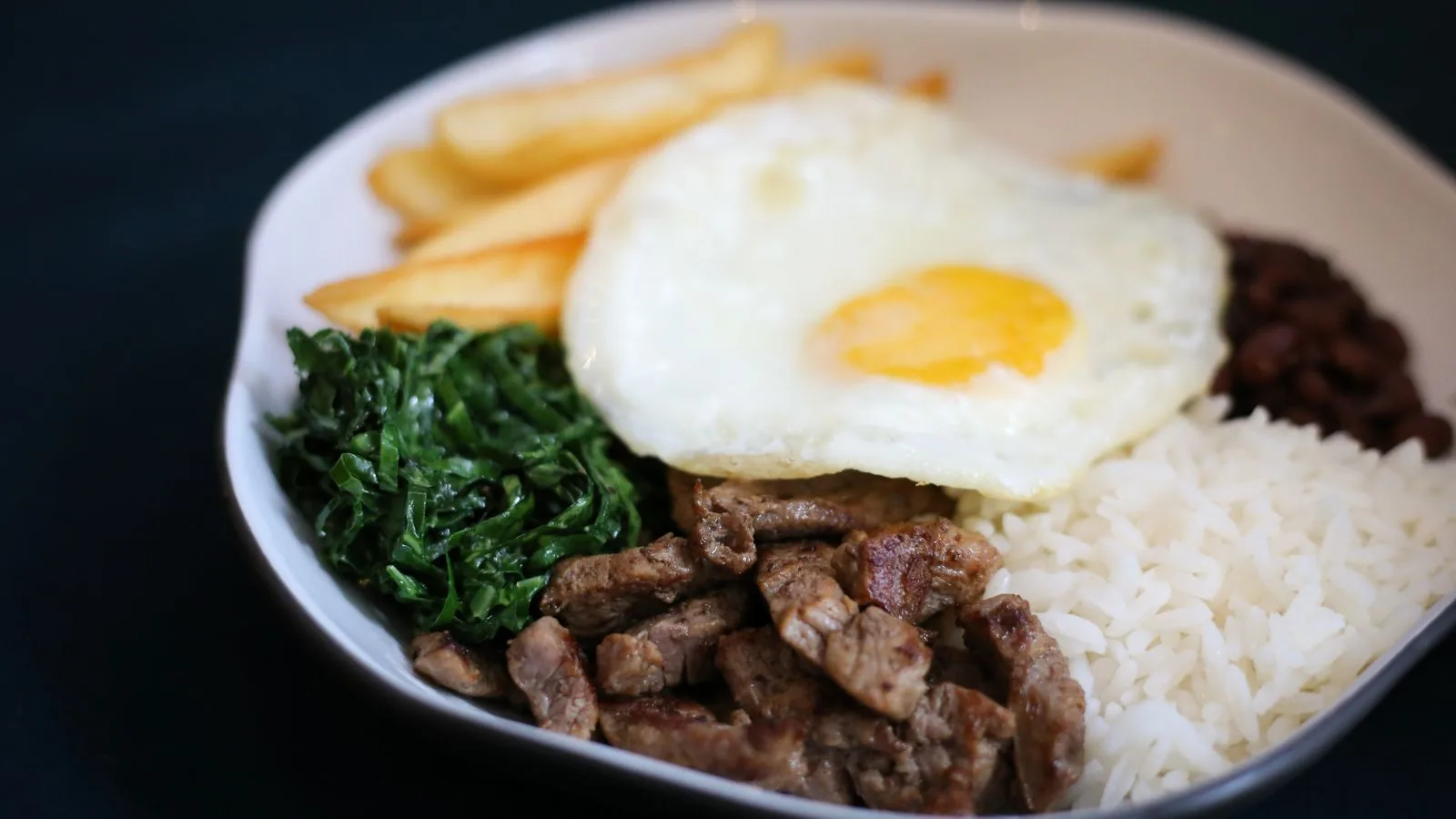 Delicious plate featuring Brazilian steak, fried egg, rice, beans, fries, and greens.