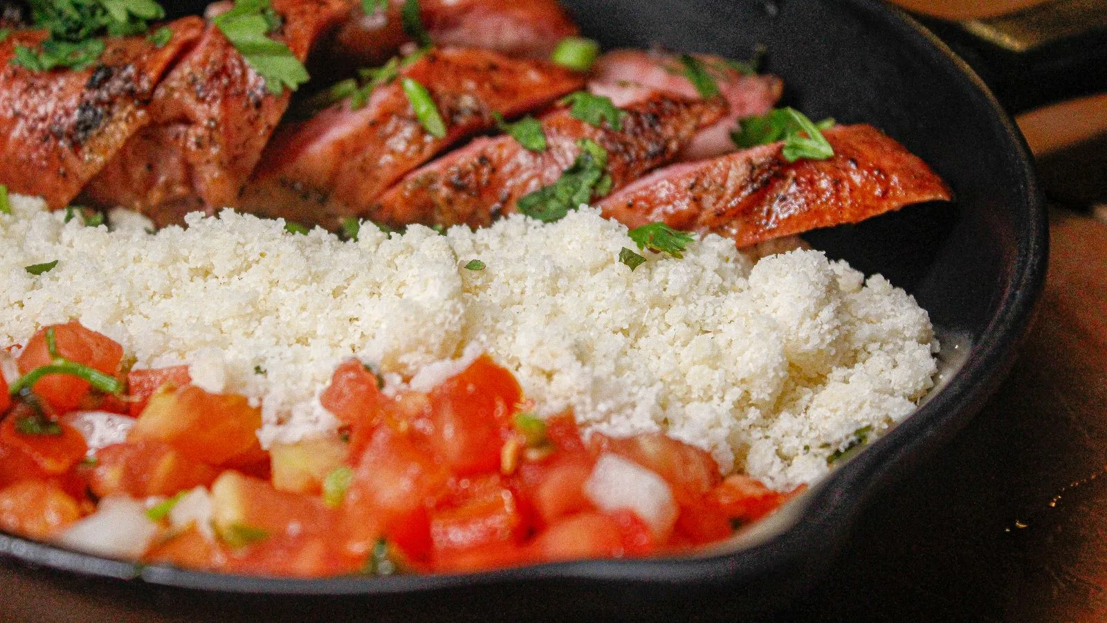 Close-up of a Brazilian dish with seasoned chicken, fresh farofa, and salsa in a skillet, capturing vibrant flavors.