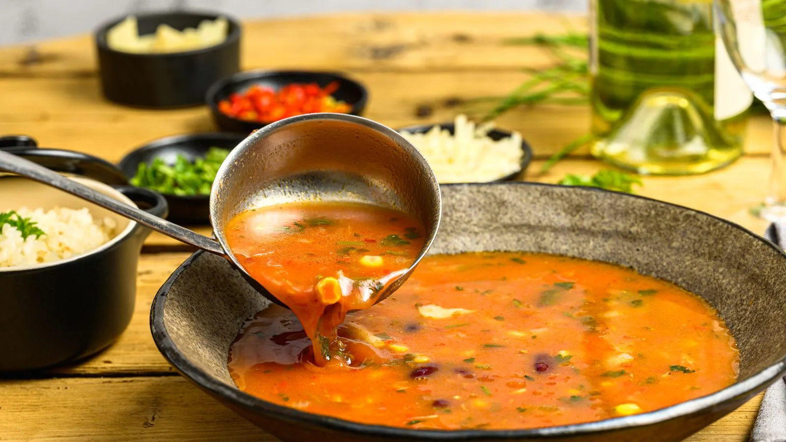 Close-up of a ladle serving vegetable soup in a rustic bowl with wine and side dishes.