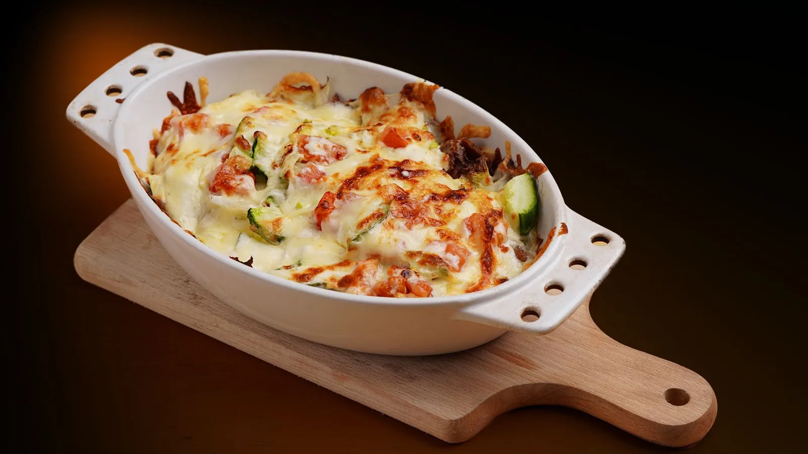 Delicious baked vegetable casserole topped with cheese in a white dish on a wooden board.