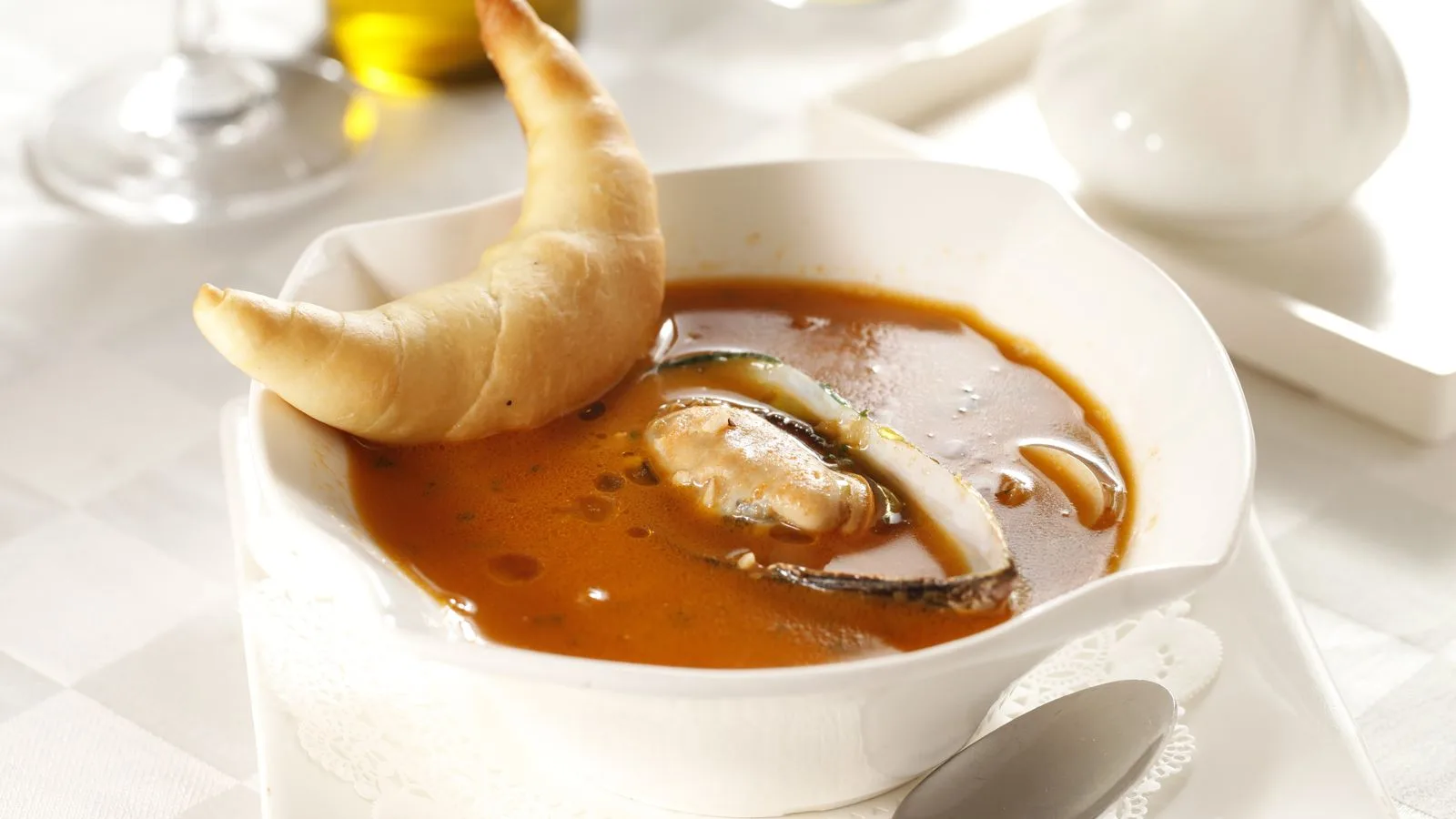 Rich tomato seafood soup with mussels served with a croissant on a white table setting.