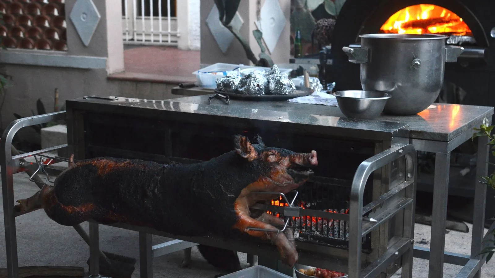 Whole smoked pig on broach near metal table with various utensils in yard of countryside house