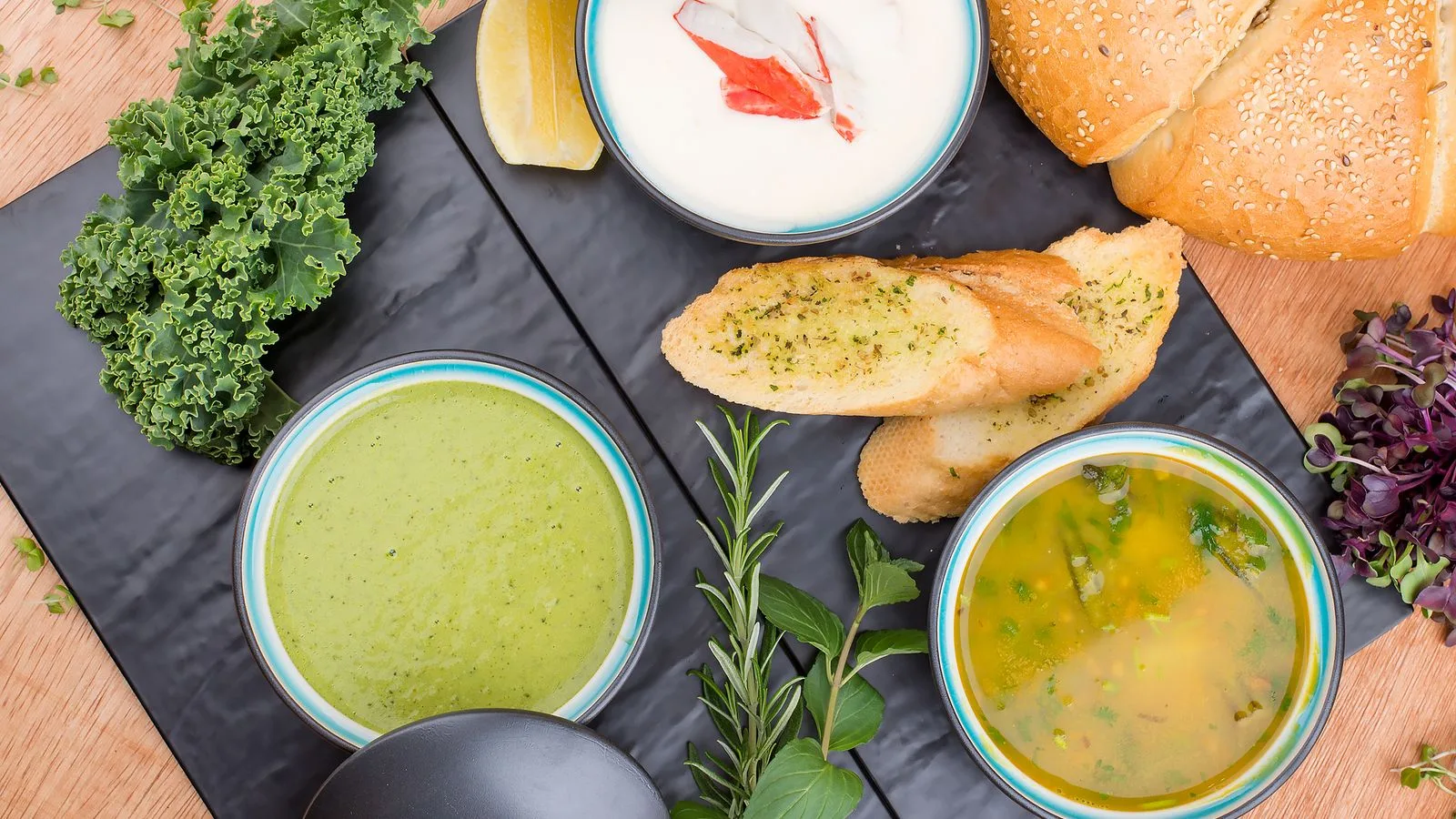 A selection of gourmet soups with fresh bread, herbs, and greens presented artfully on a wooden table.