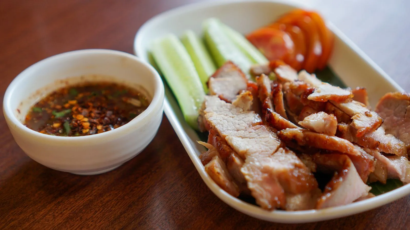 Close-up of grilled pork slices with cucumber, tomato, and dipping sauce for a flavorful meal.
