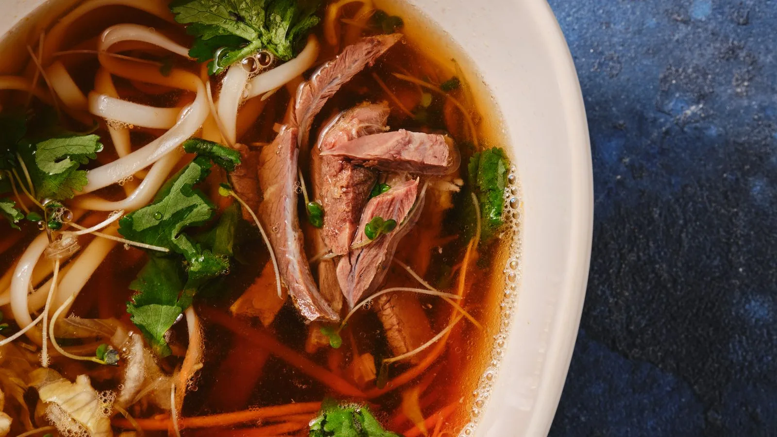 Delicious Asian beef noodle soup garnished with fresh herbs, captured in a close-up shot on a blue background.