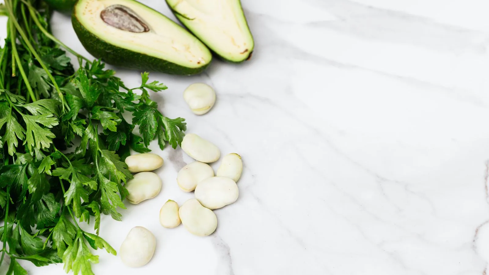 Fresh avocados, parsley, and broad beans on a white marble surface. Ideal for culinary or healthy lifestyle themes.