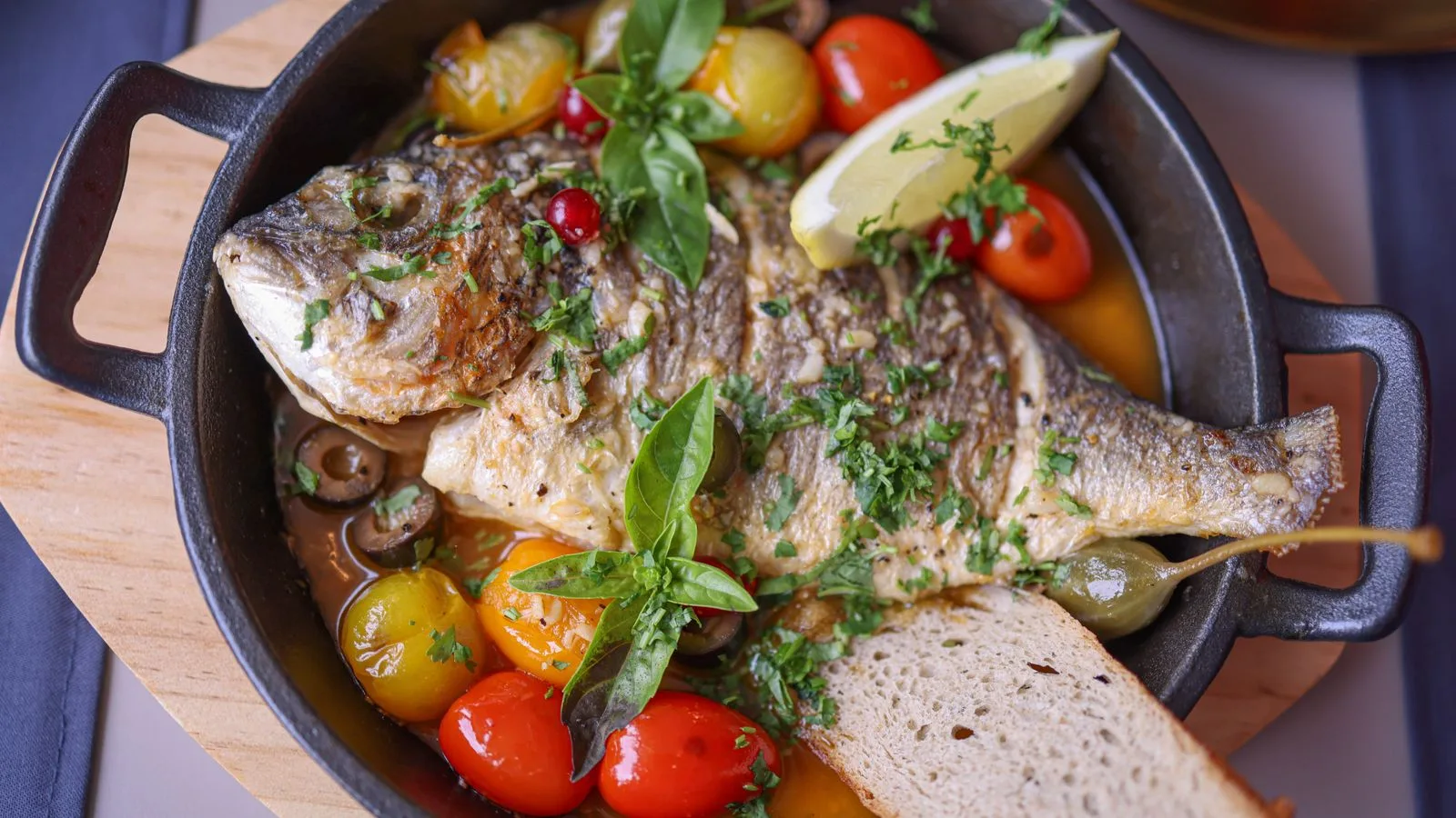 Delicious roasted fish with basil, tomatoes, olives, and lemon slice in rustic presentation.