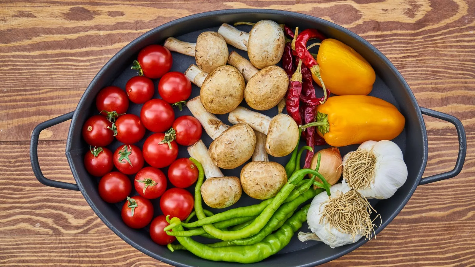 A top-down view of fresh vegetables including tomatoes, mushrooms, peppers, and garlic in a circular pan.