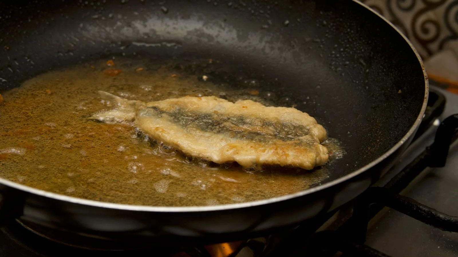 Close-up of a fish fillet frying in a pan with bubbling oil on a stove.