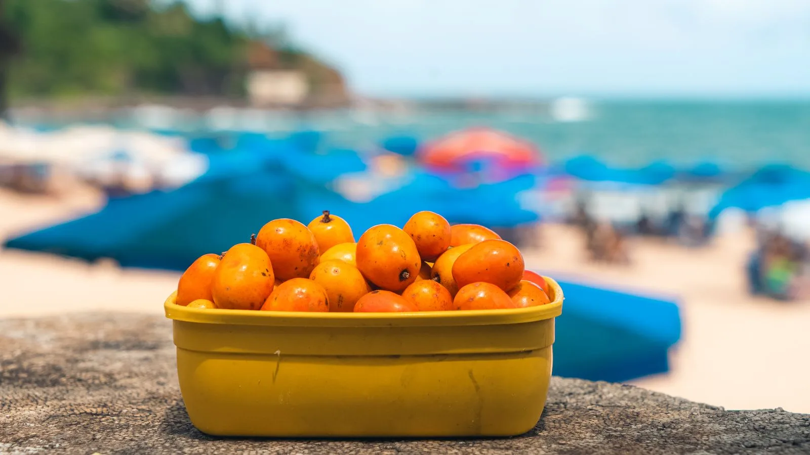 A bright yellow container of fruits sits on a beach with colorful umbrellas in the background.