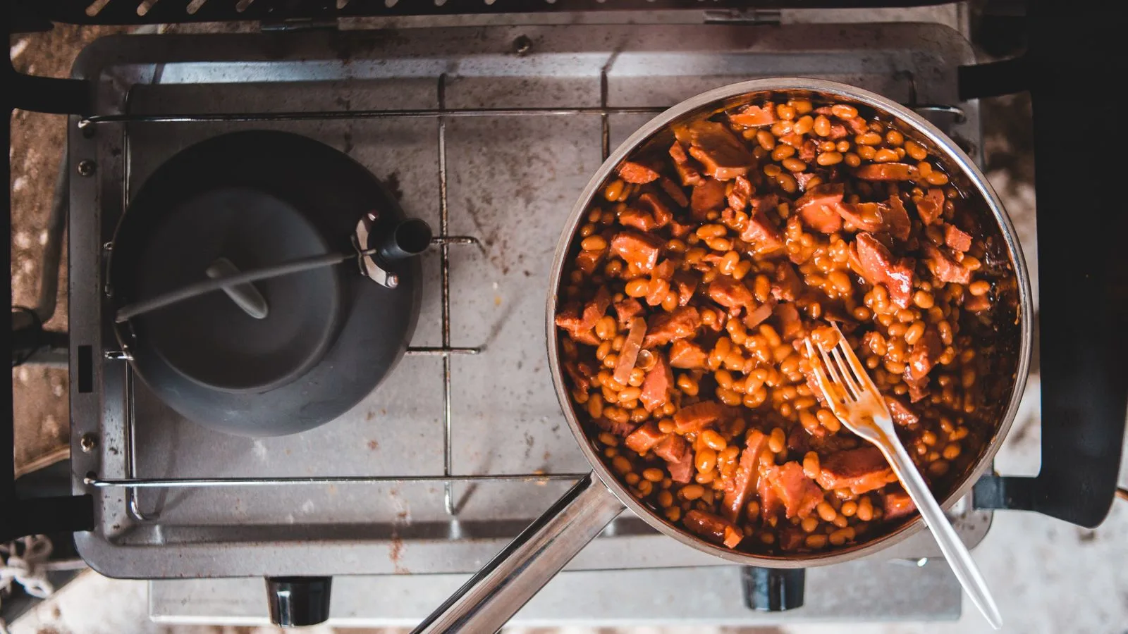 Delicious baked beans and sausage simmering on a stove in a stainless steel pan.