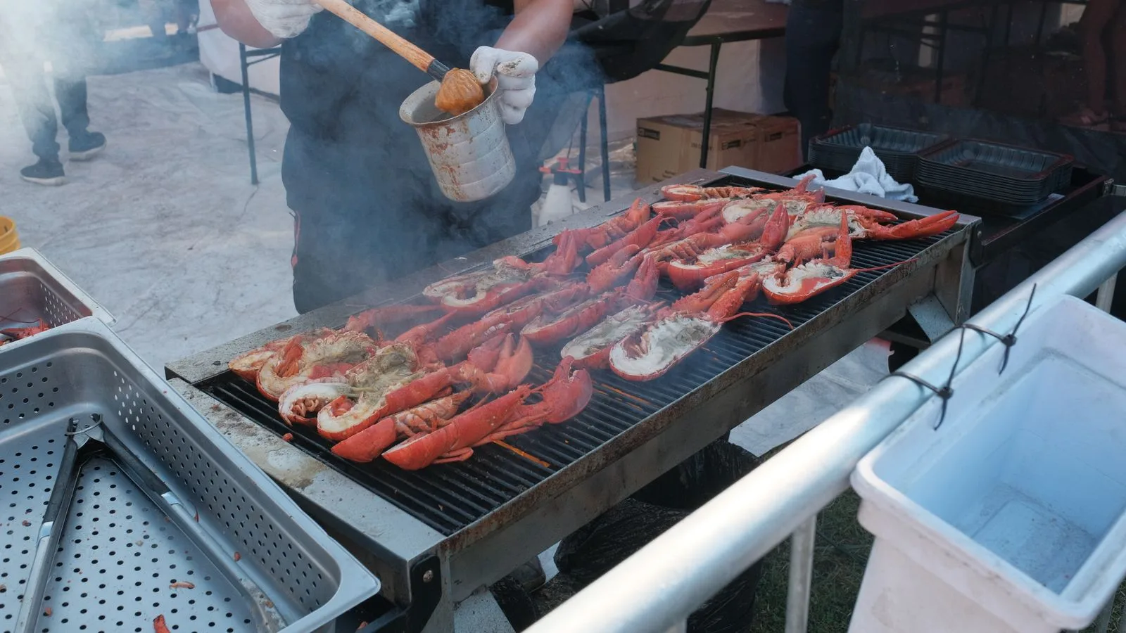 Lobsters being grilled by a man at an outdoor barbecue event with smoke rising.