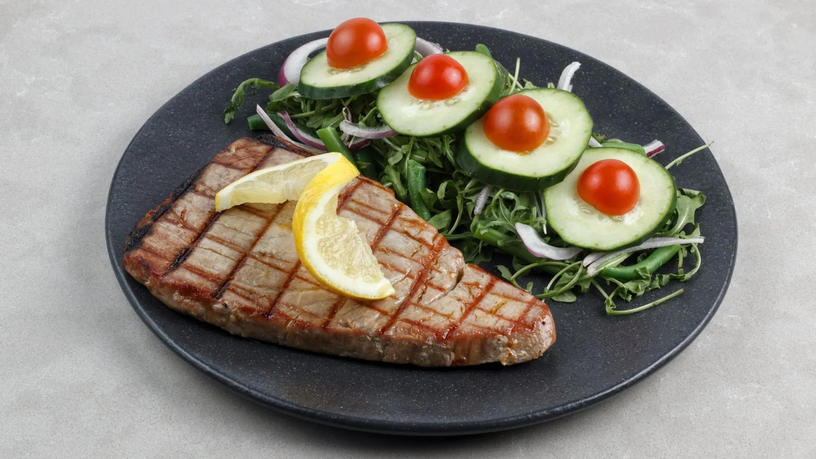 A grilled tuna steak topped with lemon slices served with a fresh cucumber and tomato salad on a dark plate.